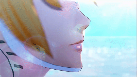 A blonde girl with short hair stands on a beach overlooking glistening blue water. She wears what appears to be a metal headband or headphones which cover her ears. Her face is framed by a white metal material that covers her entire neck and the outline of her jaw. She slowly turns to the camera.
